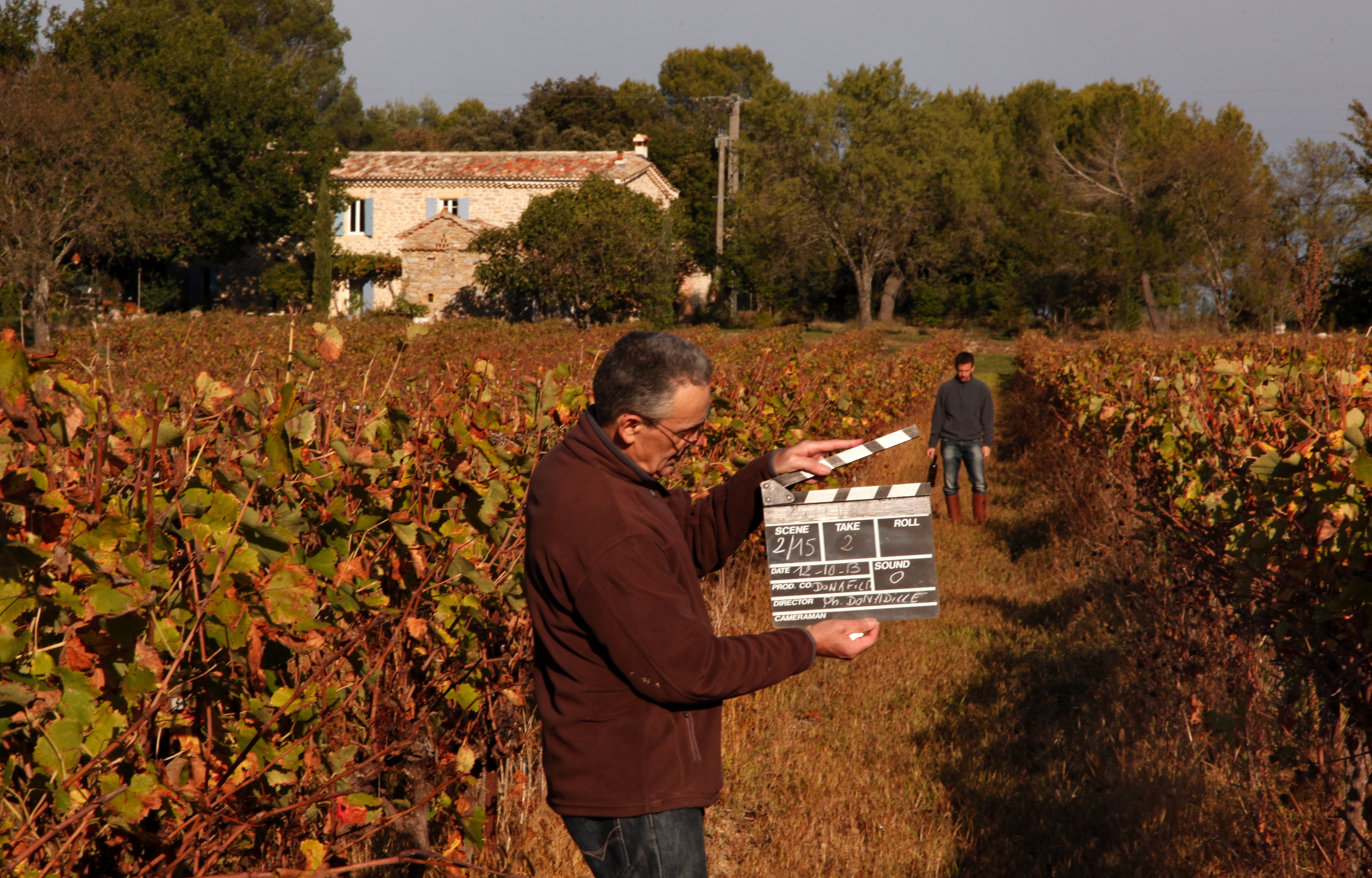 Un membre de l’équipe tient un clap dans la vigne, la demeure en pierre au loin.