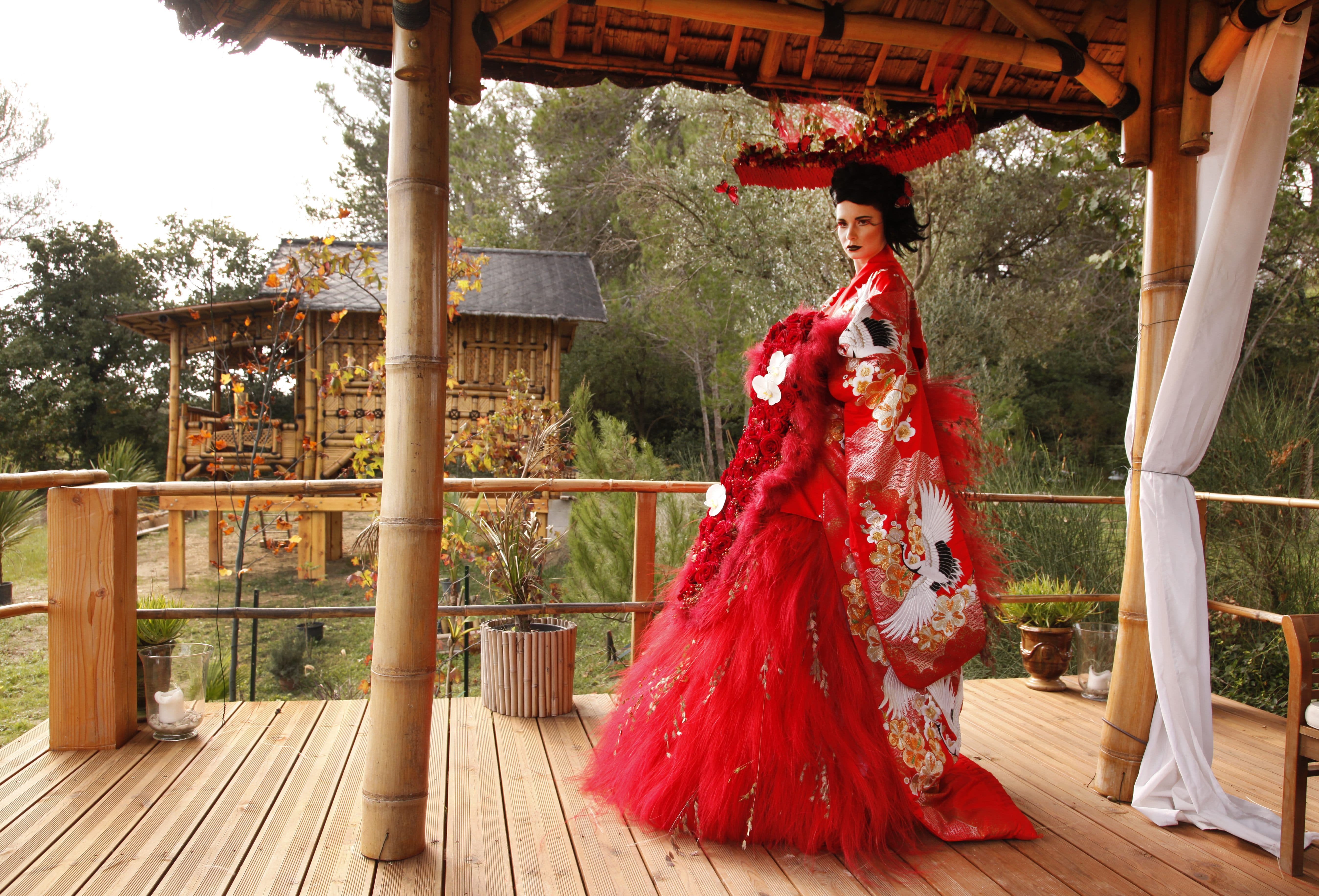 Mannequin en robe couture rouge et ombrelle sur la terrasse en teck, maison thaïe derrière.