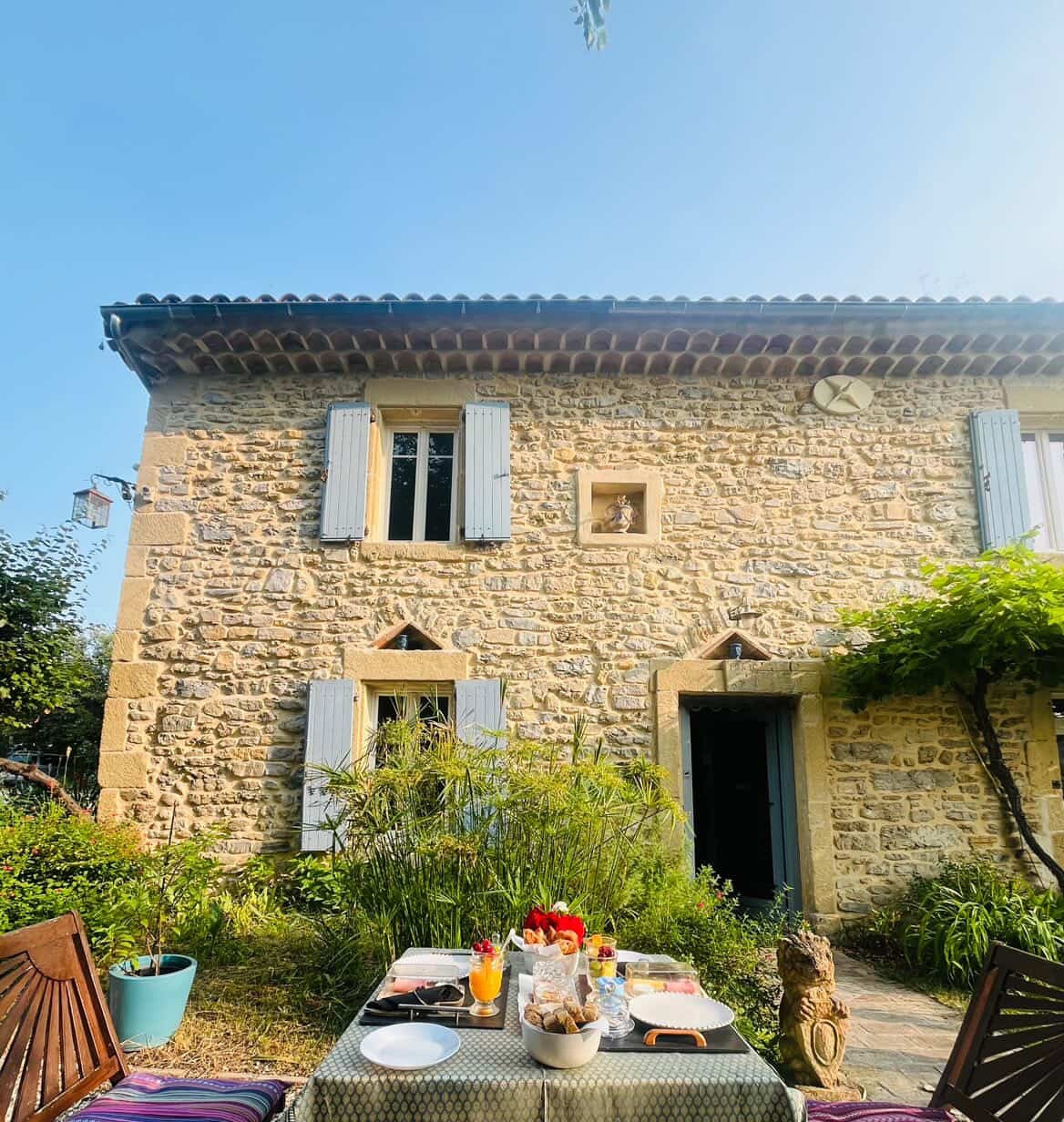 Table du petit-déjeuner dans le jardin, devant la façade en pierre du mas